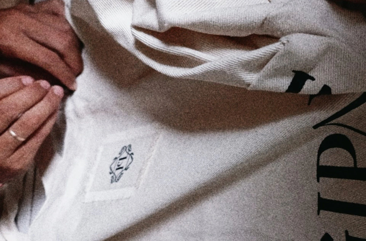 Dłonie przy jasnej torbie z czarnym nadrukiem i małą metką z monogramem.